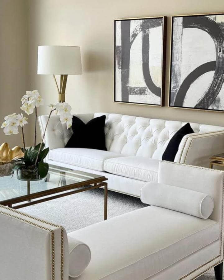 White tufted sofa and chaise with black pillow accents from @colleengahry.interiordesigner in a modern room with abstract wall art and gold lighting elements.