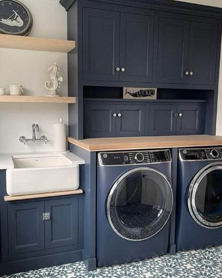 Classic navy cabinetry laundry room by @woodworkingunlimitedkitchens featuring wood countertops, white farmhouse sink, and patterned tile floor.