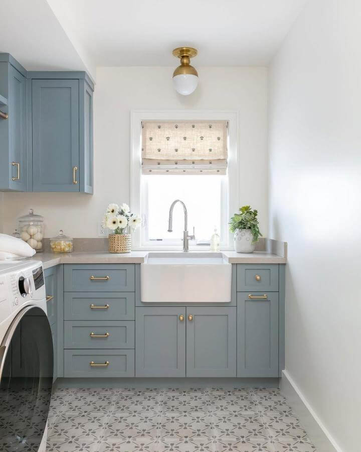Soft muted blue cabinetry laundry room from @ryangarvin featuring gold hardware and patterned floor tiles as blue laundry room ideas.