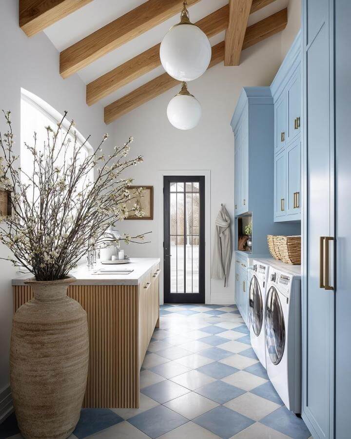 Soft blue cabinetry and cream checkerboard tile laundry room from @lifestylem_interiordesign with warm wood beams and brass details.