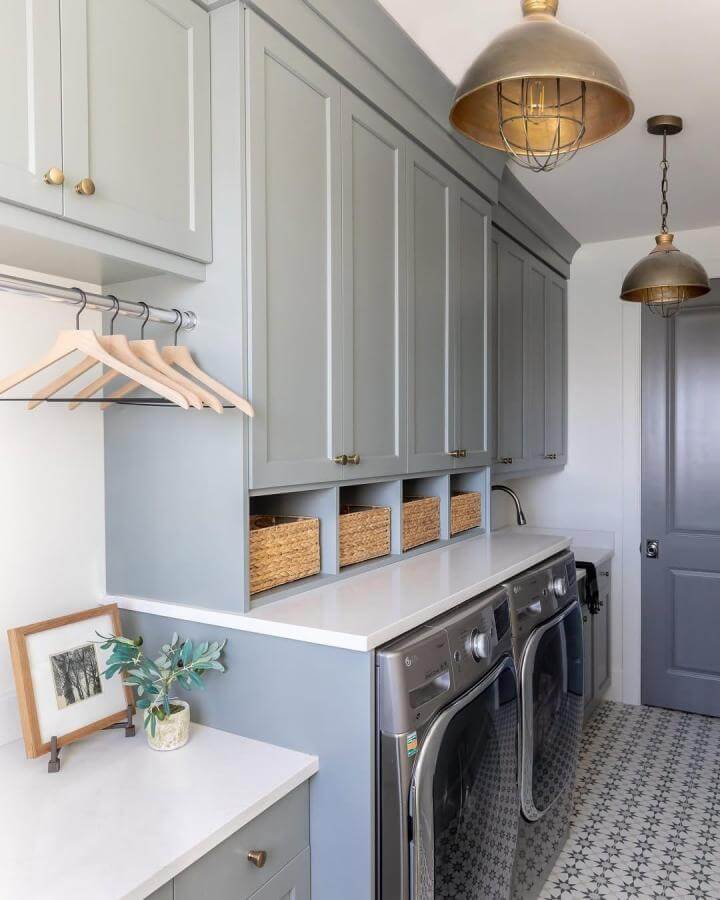 Soft muted blue cabinets and gold accents in laundry room from @lindsay_salazar_photography featuring woven baskets and patterned tile perfect for blue laundry room ideas.