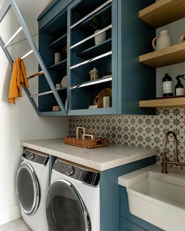Soft muted blue cabinetry in laundry space from @lindsay_salazar_photography with patterned tile backsplash and natural wood shelves.