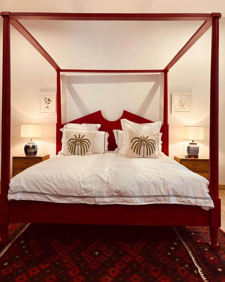 Cherry red four-poster bed with white linens and tropical embroidered pillows from @sourcingbali in a warmly lit bedroom setting.