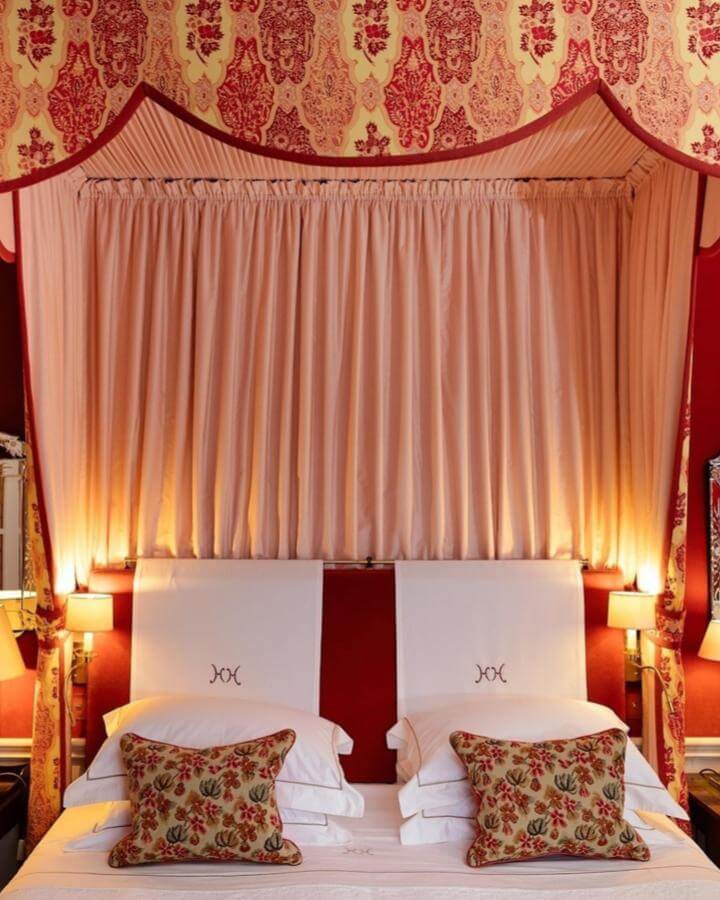 Rich red and gold damask canopy drapes with soft peach curtains and floral pillows in an elegant bedroom from @hambleton_hall.
