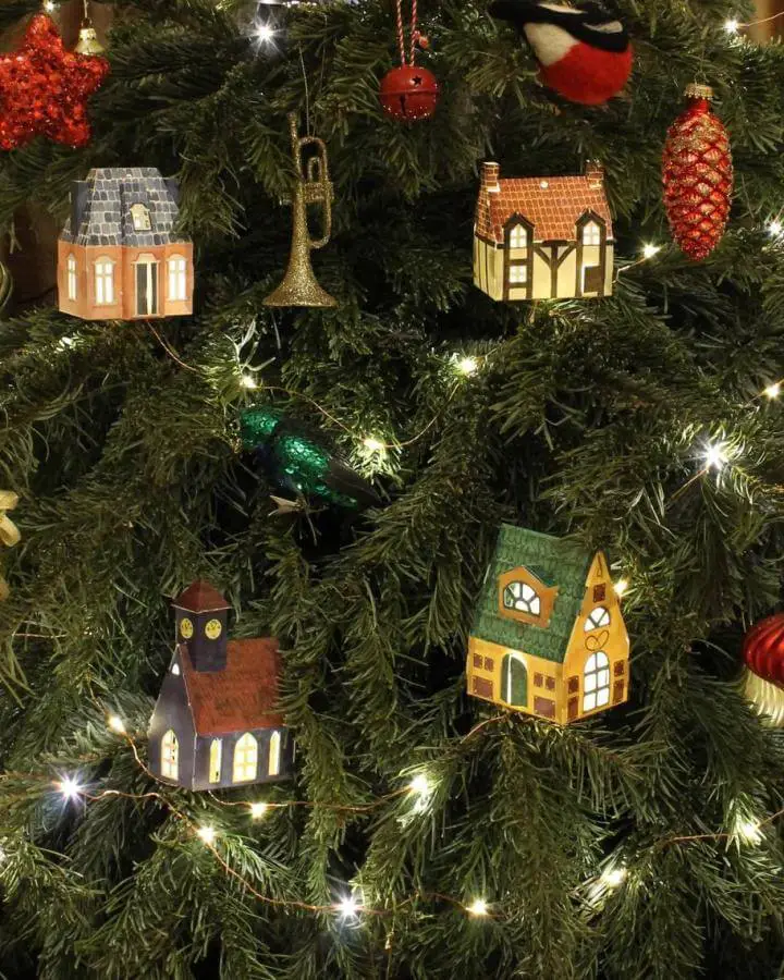 Miniature cardstock house ornaments from LittleCityLightsShop with illuminated windows hanging on evergreen tree branches.