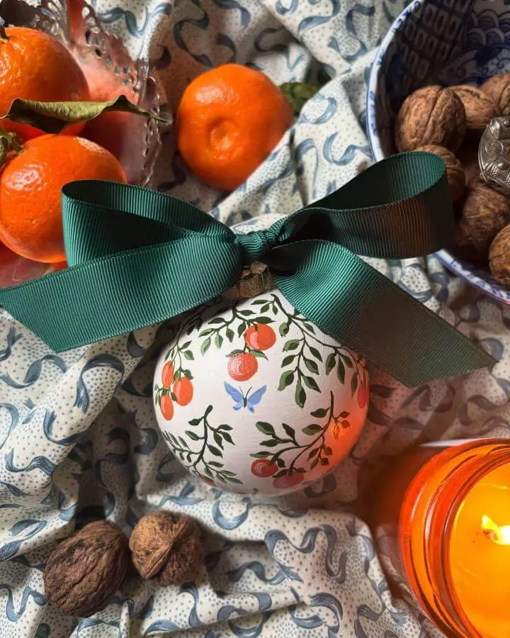 Hand-painted botanical ornament from @felicitybuchanandesigns featuring oranges, leaves, and butterfly with green ribbon.