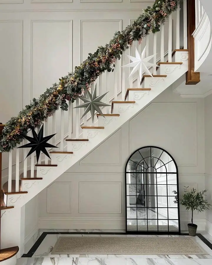 White staircase with mixed greenery garland and black paper stars from @lifeasmrsbuilder featuring warm lighting accents