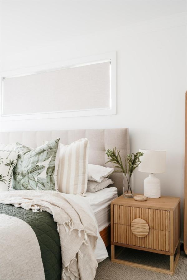 Natural wood nightstand with tropical leaf pillow from @lydiamaskiellinteriors. Neutral beige headboard with layered coastal textures.