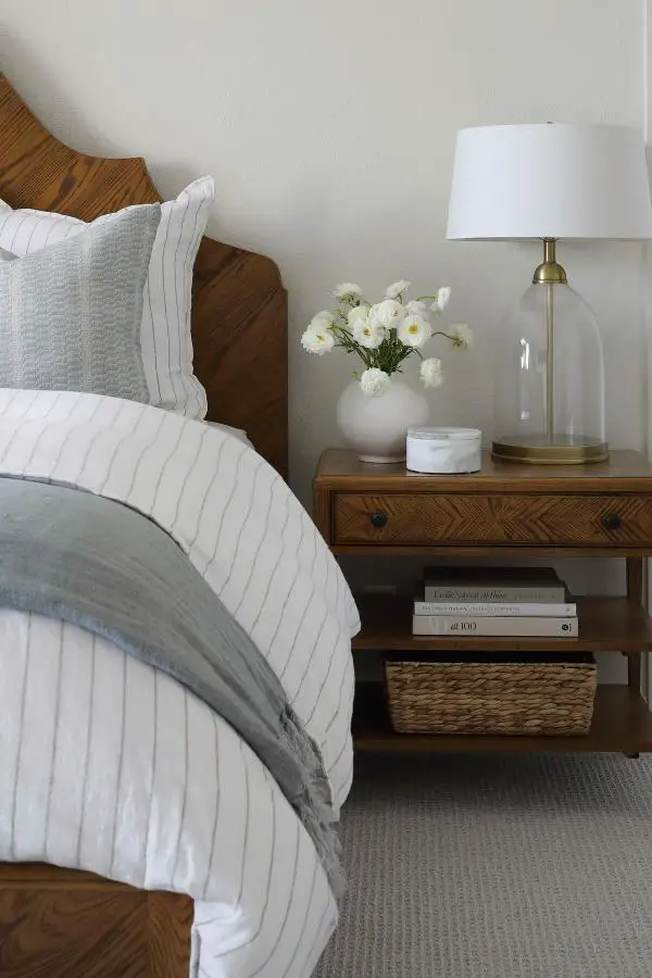 Clear glass bottle lamp from @annefrentzelinteriors. White ceramic vase with flowers on wooden nightstand beside striped bedding.