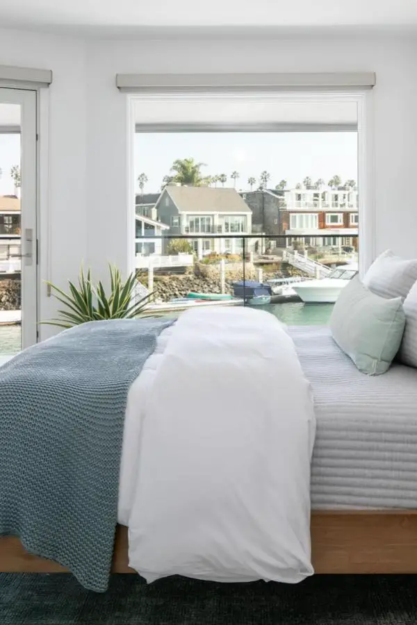 Serene waterfront bedroom from @ashleydonohuedesign. White bedding with seafoam accents overlooks marina with boats and coastal homes.