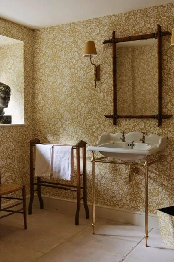 Golden damask wallpaper bathroom from @studio_squire. Brass console sink with wooden towel rack and vintage mirror.