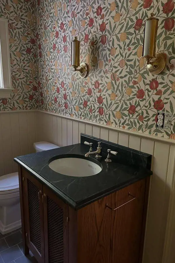 Botanical floral wallpaper bathroom from @megmcsherryinteriors. Features brass sconces and white wainscoting with dark marble vanity.