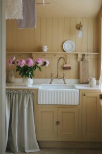 Buttery yellow kitchen with white farmhouse sink from @louiseroehome. Vintage brass faucet and striped curtain details.