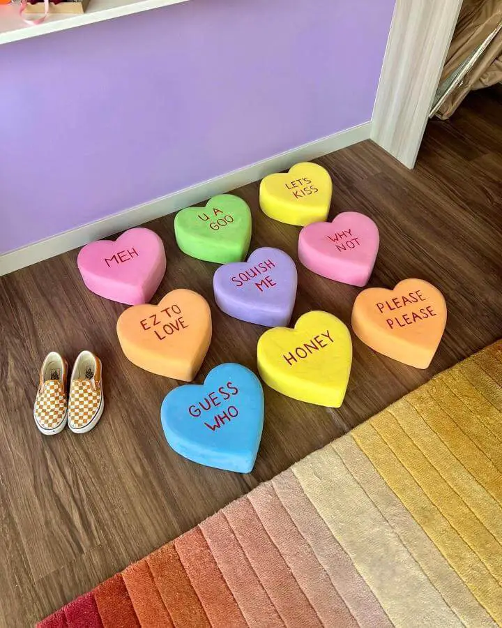 Colorful oversized heart cushions from @taybeepboop arranged on wood floor with playful text phrases.