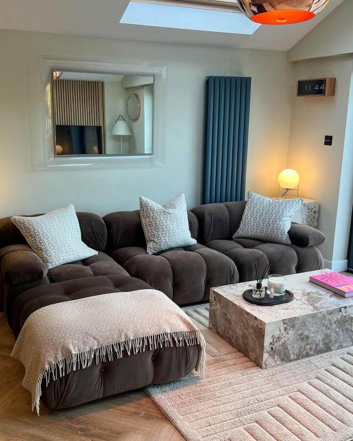 Dark brown tufted sectional with beige throw and patterned pillows in a modern living room from @sam_j_marlow. Cozy, elegant atmosphere with stone coffee table and soft rug.