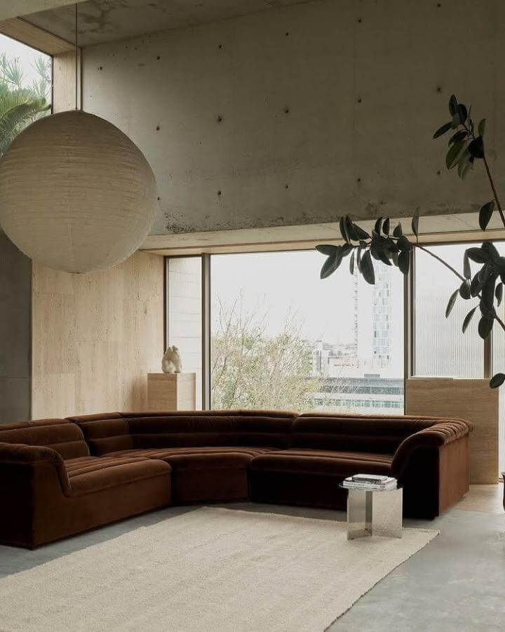 Dark brown velvet curved sectional couch in minimalist living room with concrete walls and natural light from @thedesignfiles. Cozy yet modern style.