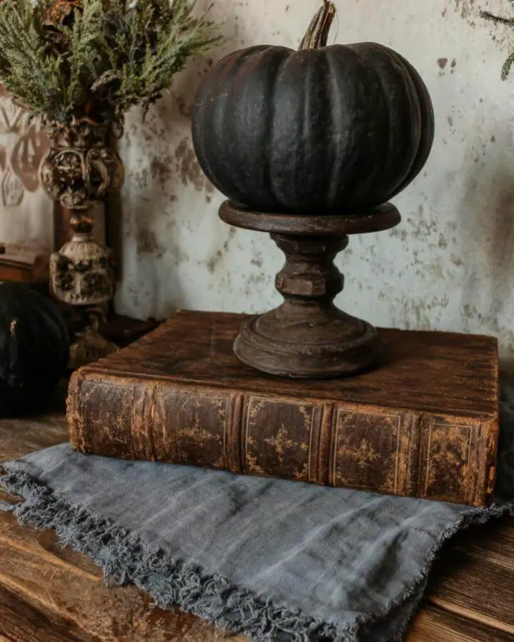 Dark green pumpkin displayed on weathered wooden pedestal with vintage books underneath. Rustic autumn styling with classical elements.