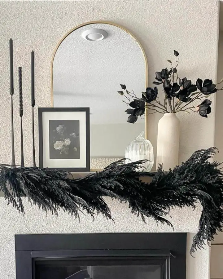 Black garland mantel display from @myneutralhomeca. Moody foliage drapes across white fireplace with candles and neutral decor.