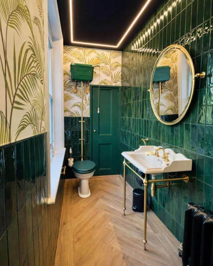 Dark glossy tile and palm wallpaper dark green powder room by @thomascrapperco with brass fixtures and wood herringbone floor