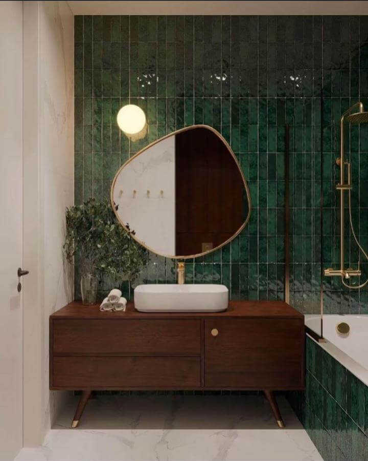 Dark green powder room with walnut vanity and stylish irregular mirror from @mayadesignstudioo. Glossy tiles and gold fixtures.
