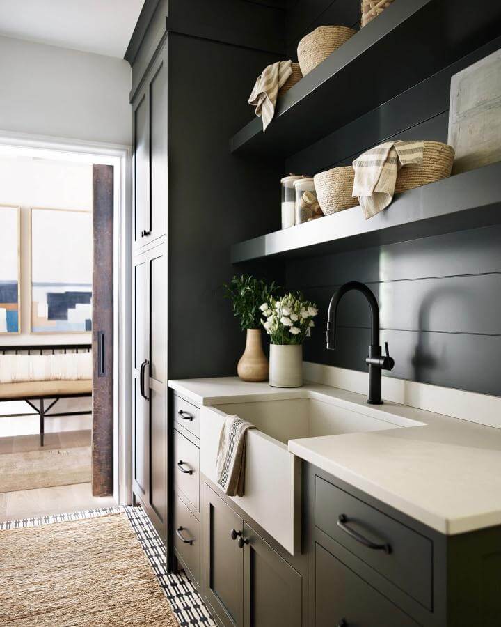 Black matte cabinets and shelves in a dark laundry room ideas space by @brookewagnerdesign featuring woven baskets and a farmhouse sink.