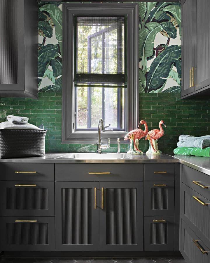 Dark gray cabinetry with forest green tile backsplash and tropical wallpaper in a stylish laundry room by @camerondesign with brass hardware and playful decor.