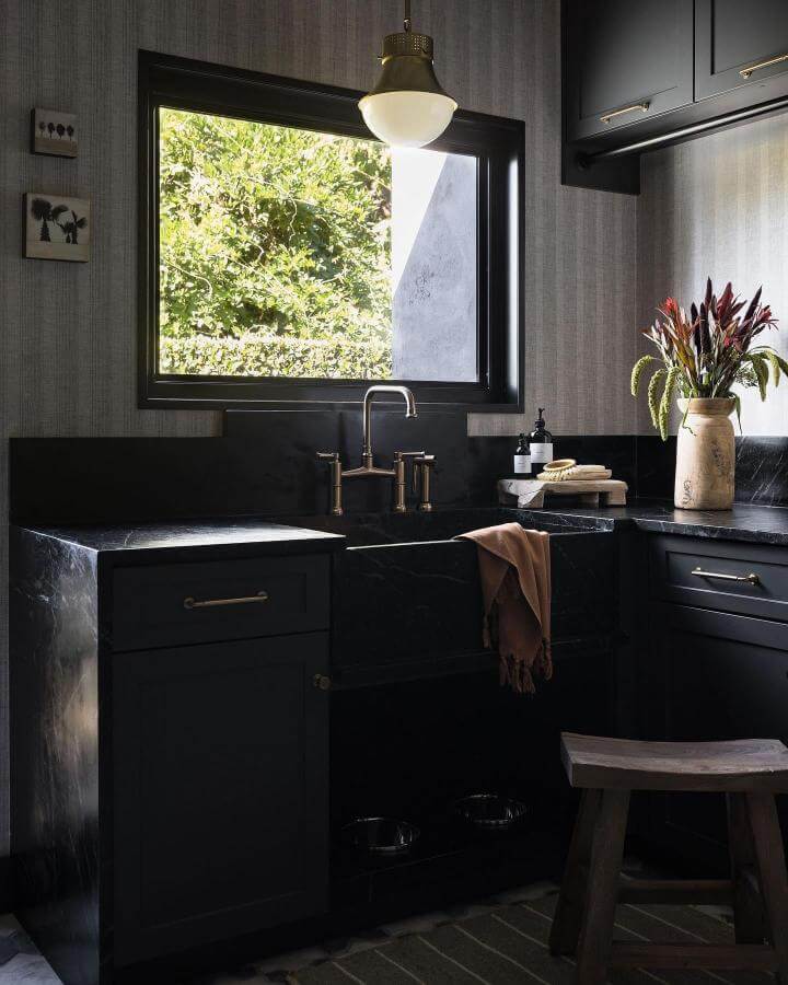 Black marble laundry sink with brass fixtures and charcoal cabinetry by @michevictoriadesign in a moody elegant room with natural light.