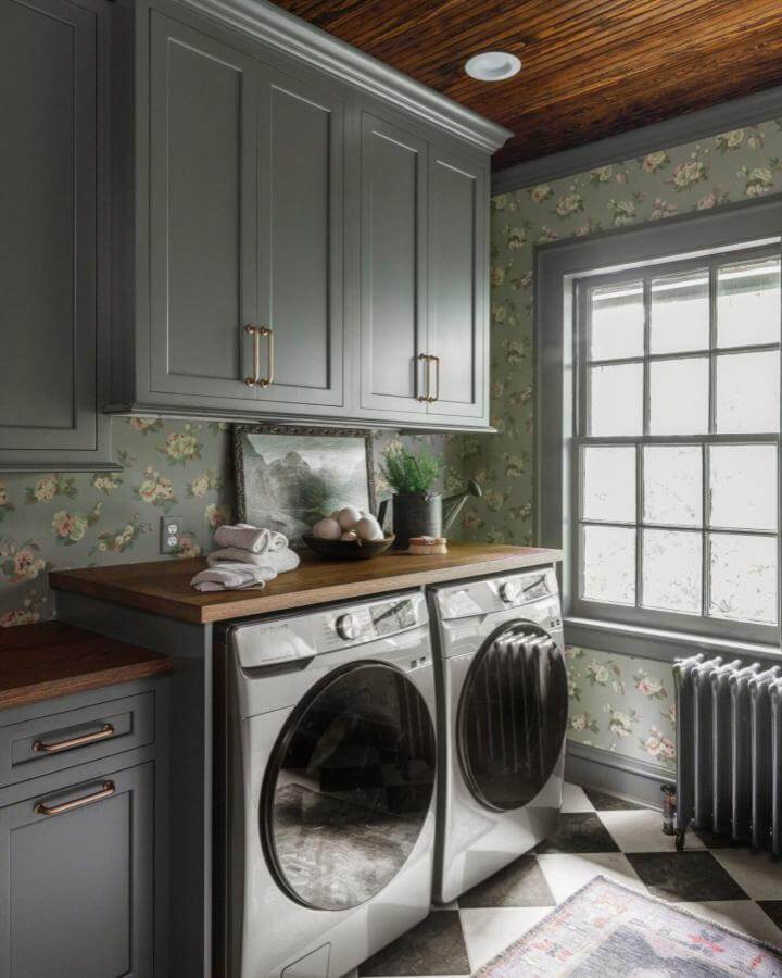 Dark gray cabinetry laundry room with floral wallpaper and wood countertop from @karrbick_design showing moody vintage style and cozy charm