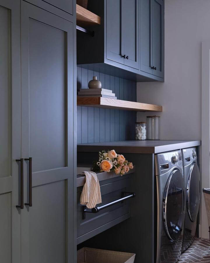 Dark blue matte cabinets laundry room with wood shelves and black hardware from @gatherhouse_design. Cozy modern space with natural accents and flowers.