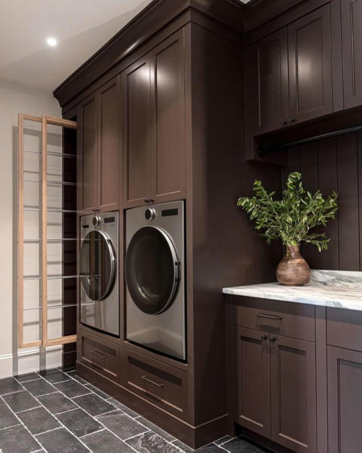 Dark espresso cabinetry and marble countertop laundry room from @verandaestatehomes. Clean lines with built-in washer dryer and wood drying rack for dark laundry room ideas.