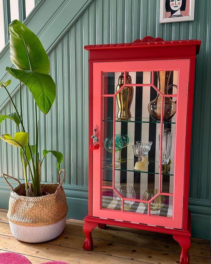 Bright coral vintage-style drinks cabinet with glass shelves and bold black and white striped background from @little_edwardian_semi showcasing unique glassware display diy drinks cabinet ideas.