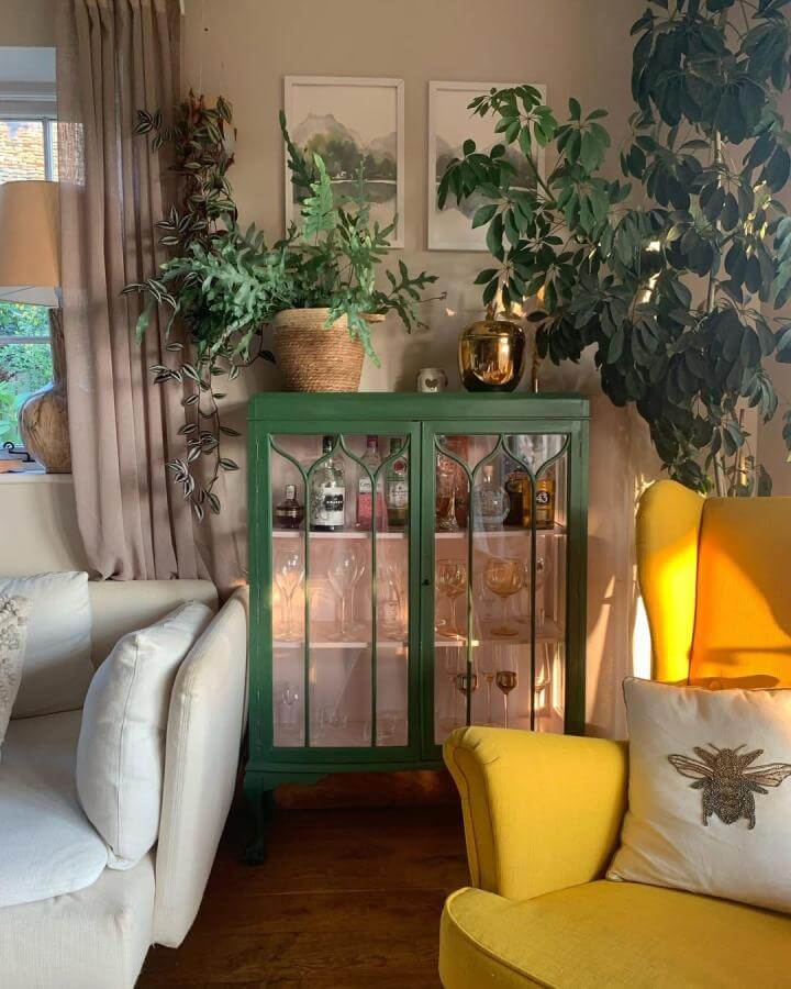 Green upcycled drinks cabinet with glass doors and pink interior in cozy living room by @what_a_view_cottage, surrounded by plants and colorful furniture.