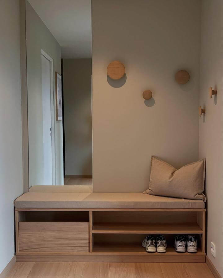 Light wood bench with beige cushion and hidden shoe shelves from @nordisk.heim in a calm minimalist entry nook with wooden wall hooks.