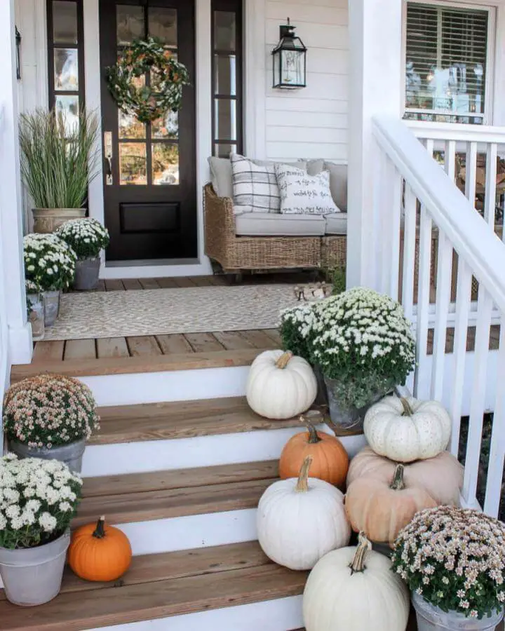 Wooden porch steps with scattered cream and orange pumpkins plus white mums in galvanized planters. Natural autumn styling.