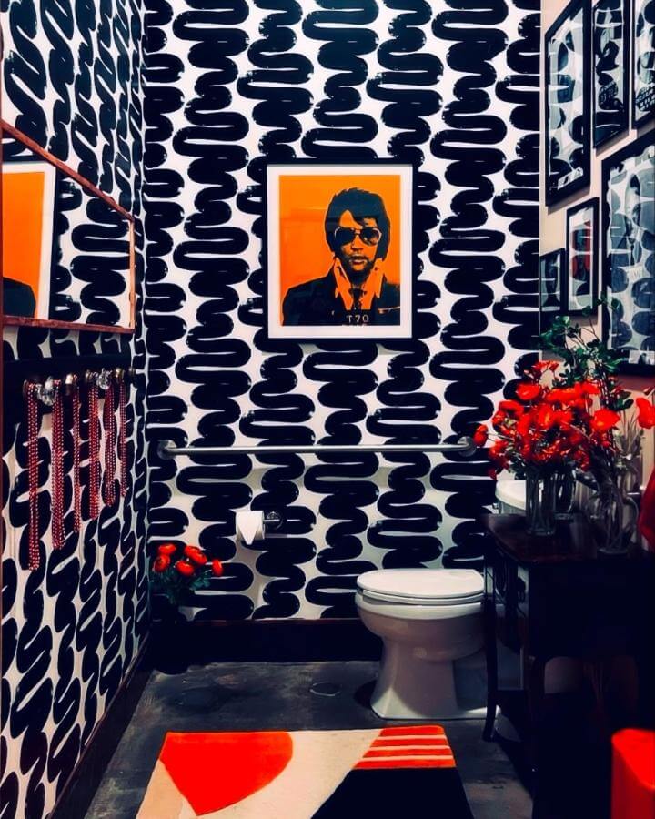 Black and white abstract patterned wallpaper bathroom with bright orange art and floral accents from @innattheoldjail funky bathroom ideas vibrant and unique design.