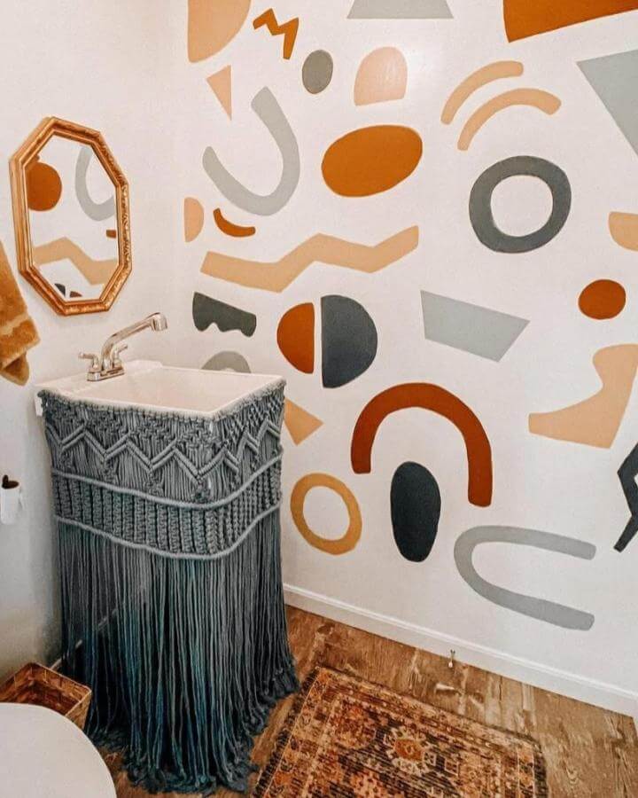 Abstract geometric wall art with earthy tones and a gray macramé sink skirt in a funky bathroom by @coda.co. Fresh and playful style.