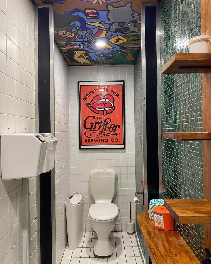 Cartoon mural ceiling in a small bathroom with green mosaic tile and wooden shelves from @reviewsfromtheloo funky bathroom ideas playful and colorful.