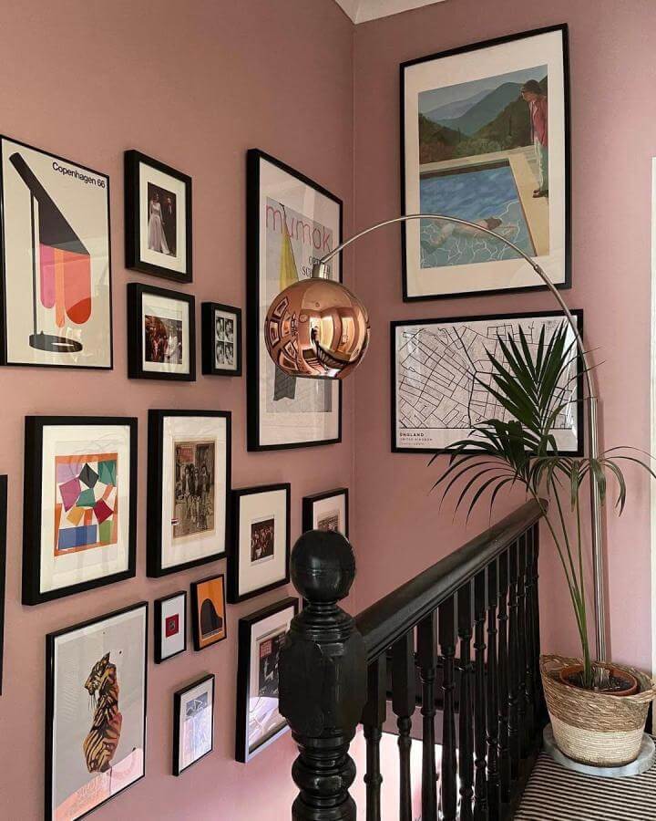 Soft pink walls with diverse black picture frames and copper lamp from @emeraldterrace bringing warmth and vibrant personality to a stairwell.