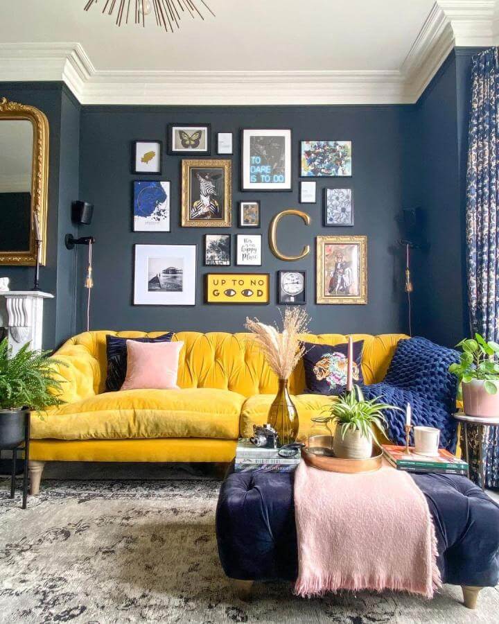 Dark navy gallery wall with mixed art and gold accents above mustard sofa from @lauracavehome showcasing layered textures and vibrant contrasts.