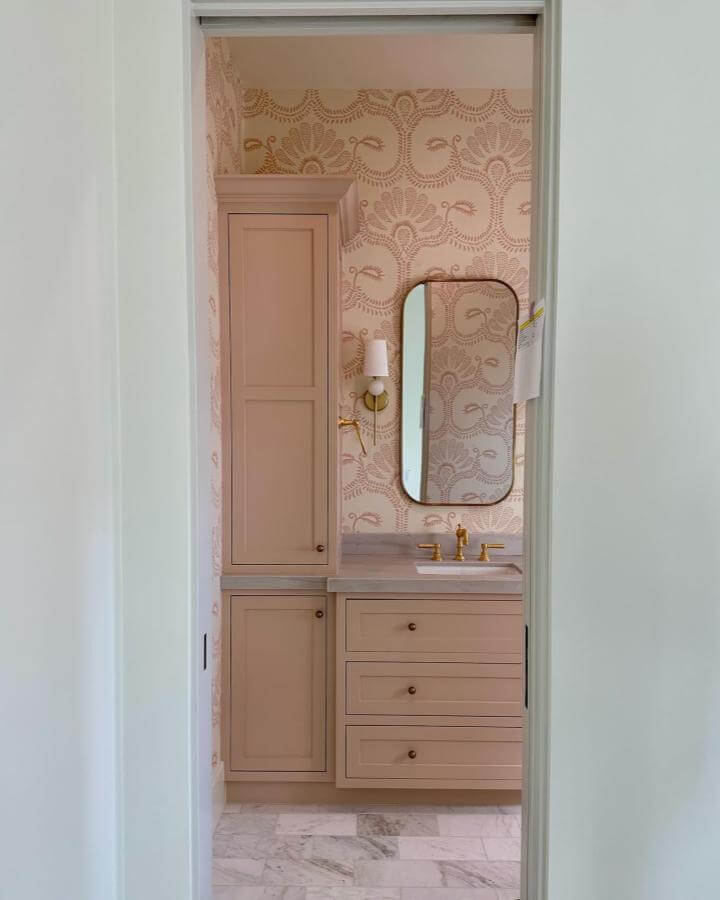 Soft blush cabinetry bathroom with gold fixtures and patterned wallpaper from @strandemo_home showcasing girly bathroom ideas. Elegant and cozy design.