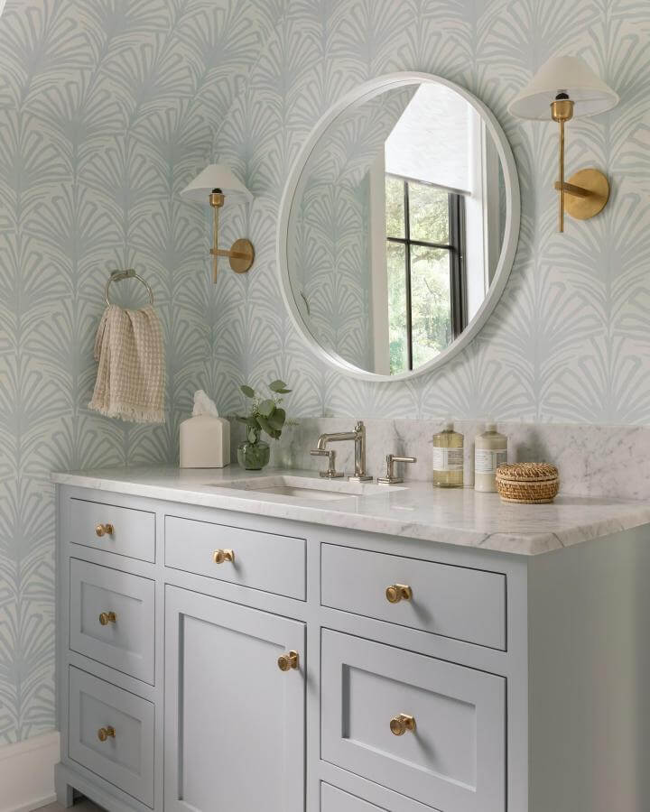 Soft blue vanity with gold knobs and rounded mirror in girly bathroom ideas by @wendymaurodesign. Elegant wallpaper and brass sconces brighten space.
