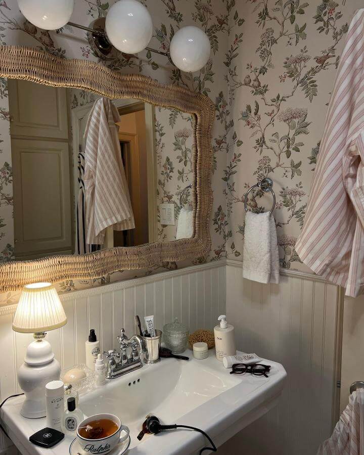 Natural wicker framed mirror and floral wallpaper in cozy bathroom from @addison_gunnnn showing girly bathroom ideas with soft pink accents and warm lighting.