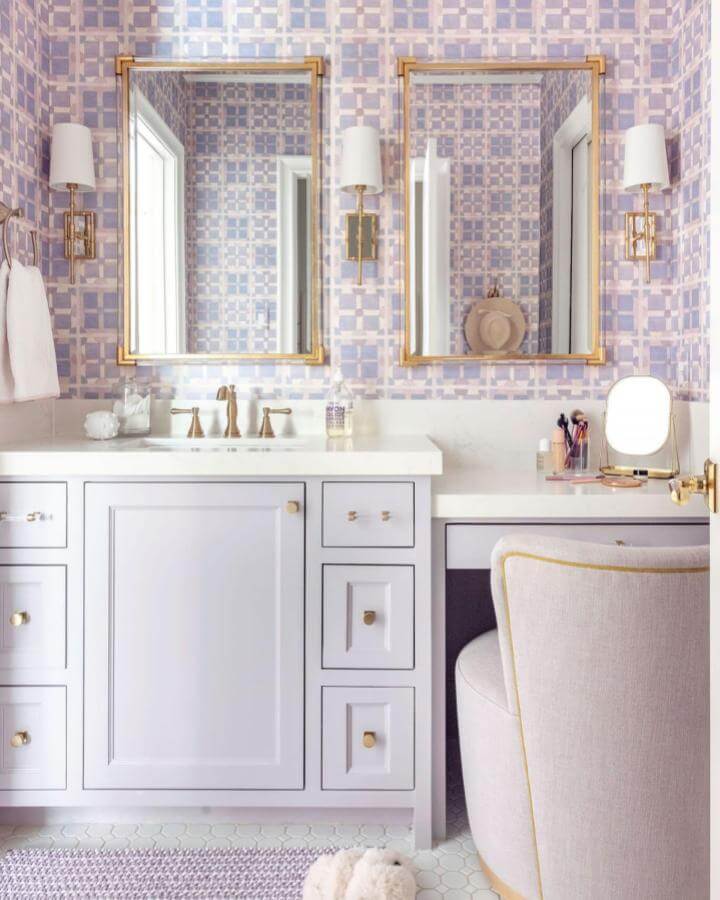 Lavender and gold bathroom vanity with patterned wallpaper from @oystercreekstudios showcasing elegant girly bathroom ideas. Plush chair and dual mirrors.
