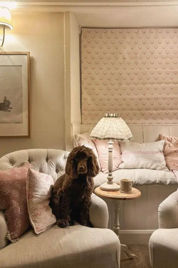 Cream table lamp from @heartsandglitter_ casting warm light over beige sectional with pink cushions and dog.