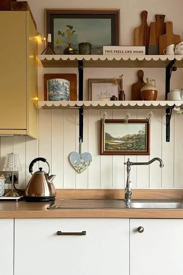 Cream kitchen with scalloped shelving from @thewalmsleyhomestead. Vintage pottery and framed landscapes create cozy cottage atmosphere.