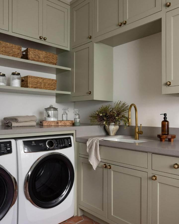 Soft sage green cabinetry and brass faucet in a green laundry room ideas setup by @stephanie___russo with wicker baskets and white appliances.