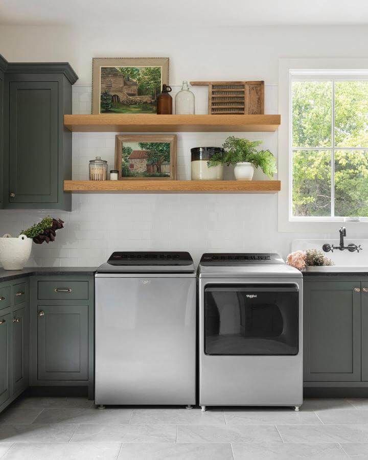 Deep forest green cabinets with natural wood floating shelves in a laundry room from @peachandpineinteriors showcasing green laundry room ideas in a cozy, stylish setup.