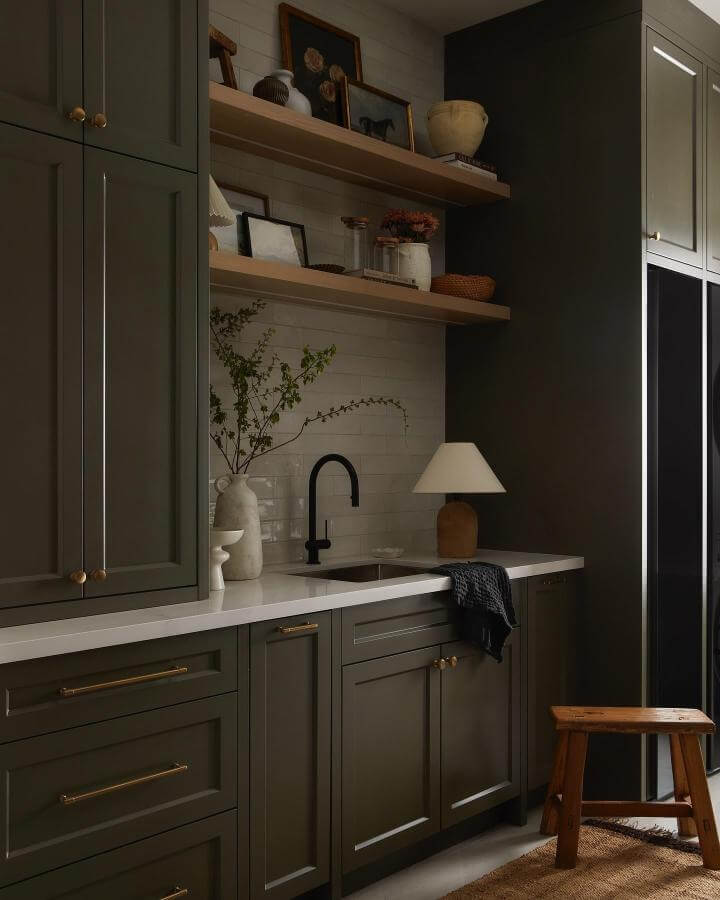 Olive green cabinetry laundry room from @lomillerphoto with brass hardware and natural wood shelves creating warm green laundry room ideas ambiance