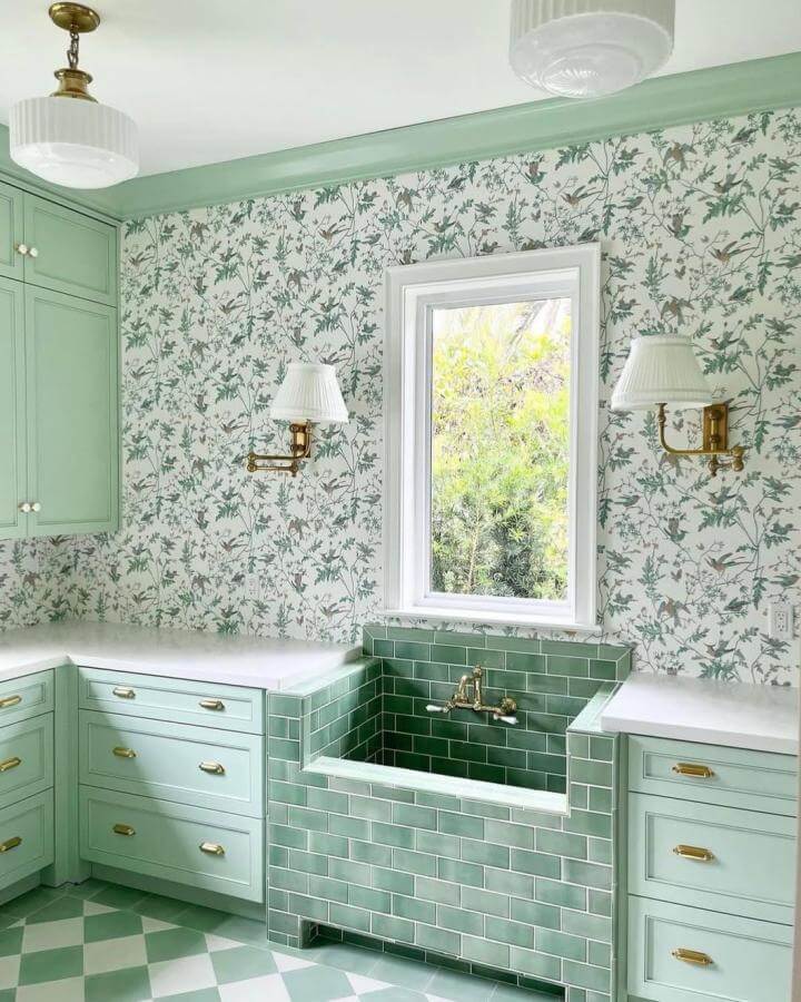 Mint green subway tile laundry sink and soft green cabinetry with floral wallpaper and brass fixtures from @karamillerinteriors showcasing green laundry room ideas. Light and fresh vibe.