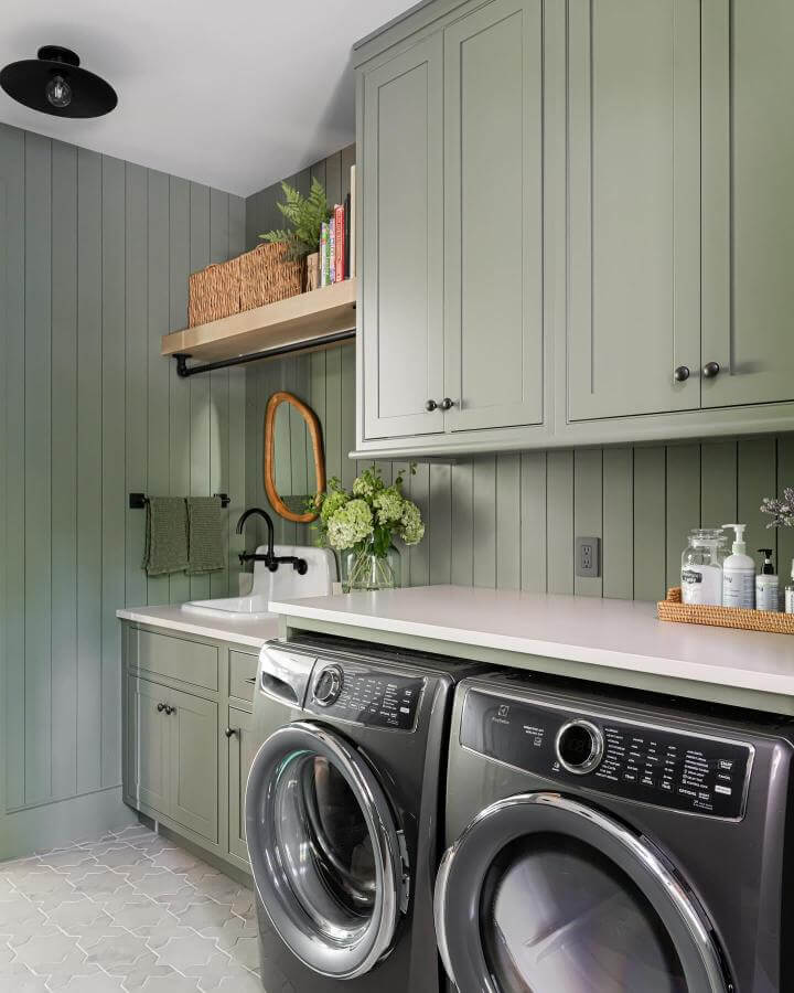 Soft muted sage green cabinetry and paneling laundry room from @lizhackettinteriors with black fixtures and natural accents green laundry room ideas.